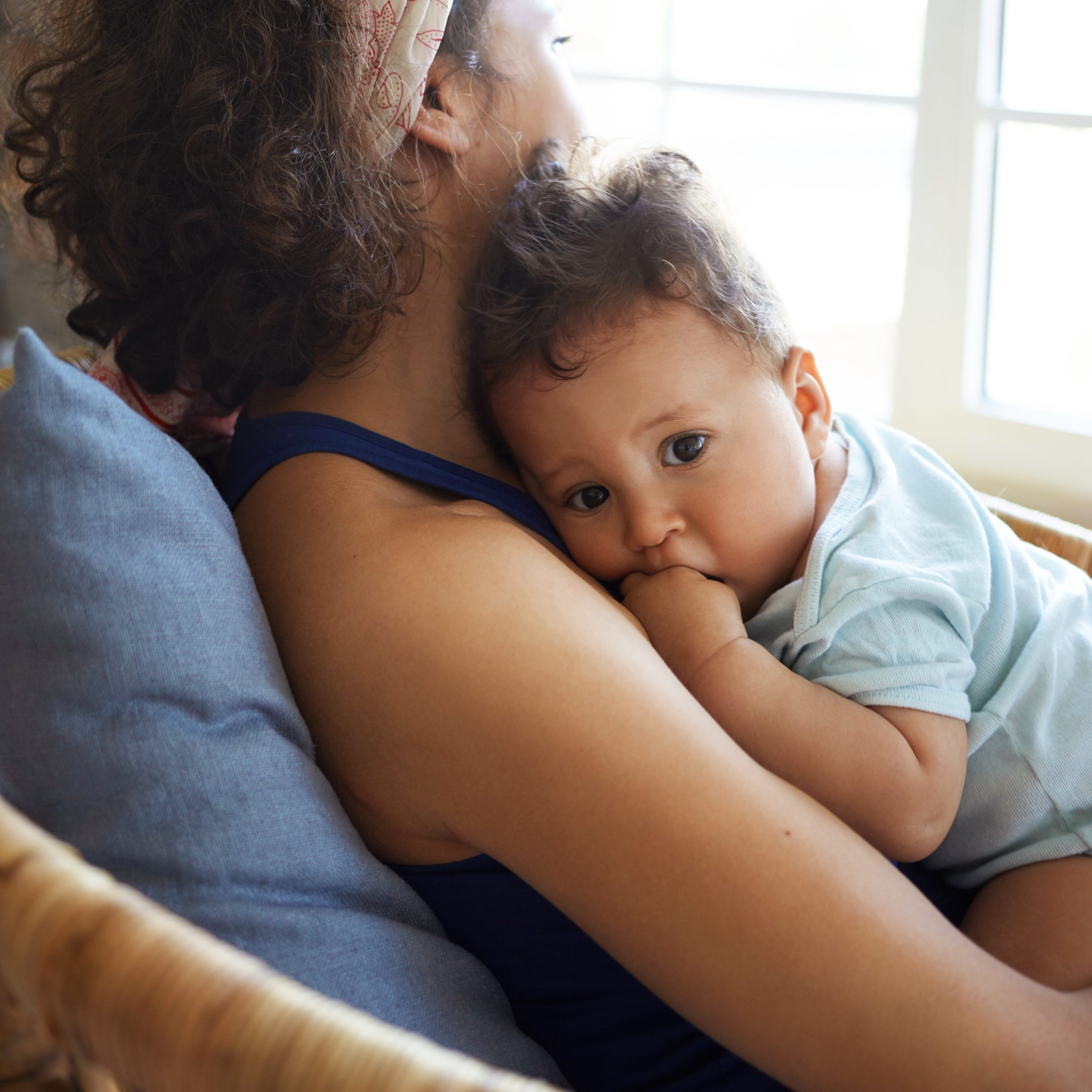 Rear view of young dark skinned female sitting by window in chair, nursing nine month old toddler. Mixed race mother soothing her infant son to sleep. Childcare, family and motherhood concept